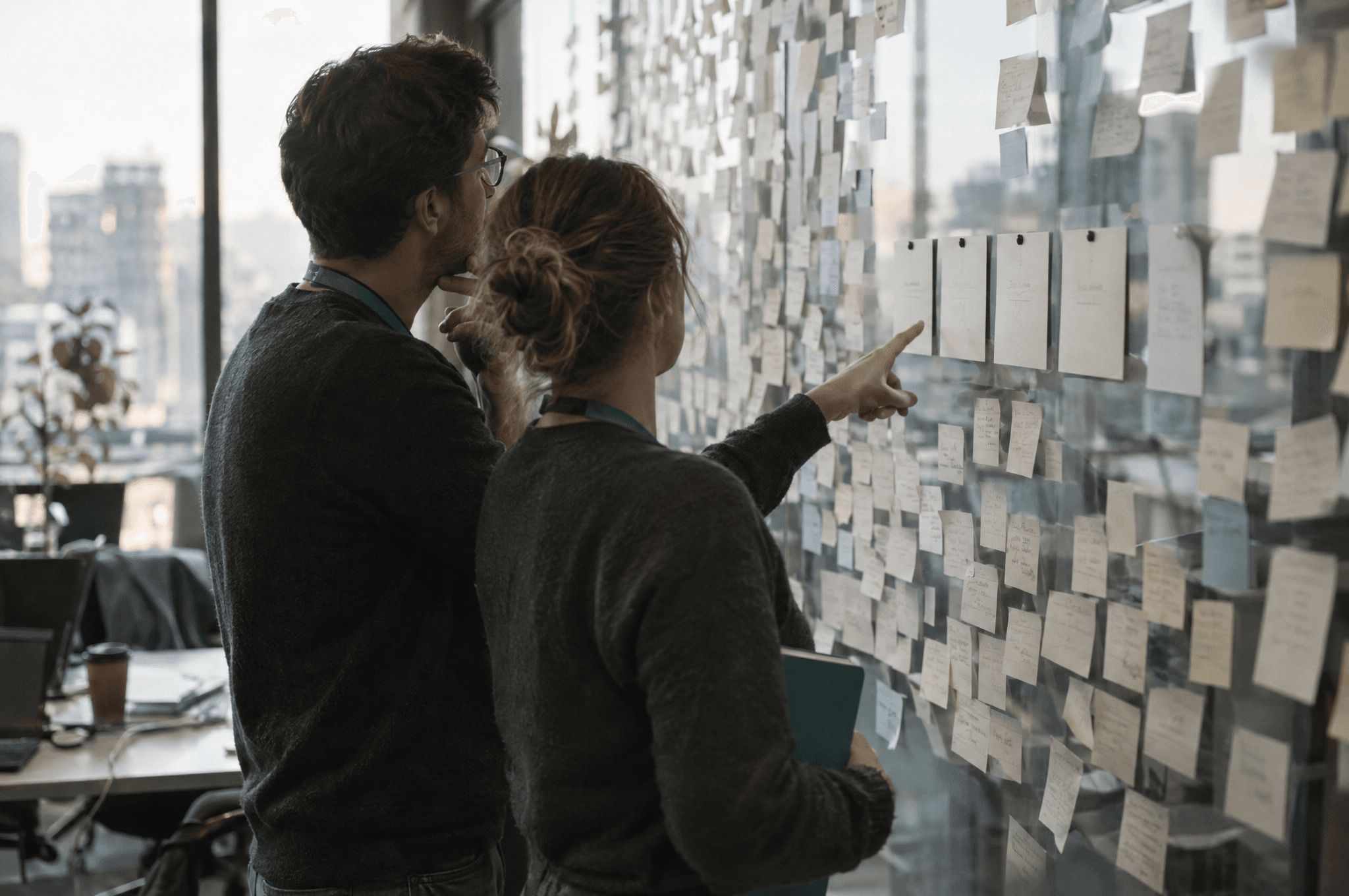Two colleagues at a glass wall covered in hundreds of sticky notes, one pointing at a row of five white cards pinned at eye level.